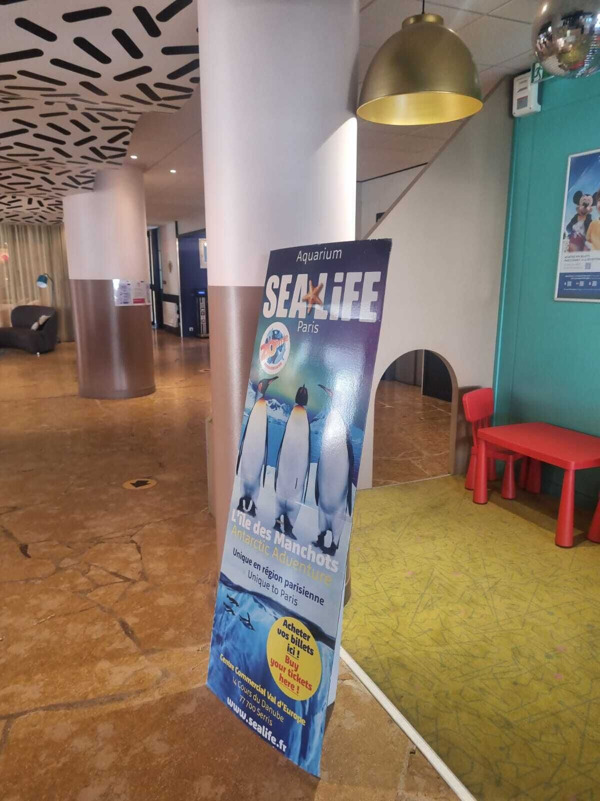 A poster for 'Aquarium SEALIFE Paris' featuring penguins, located in a lobby with a modern design, including a patterned ceiling and a red bench.