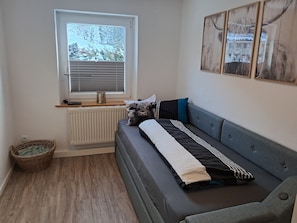 Een kamer met een grijze bank, een raam met zonwering, een radiator, een rieten mand en een ingelijst kunstwerk aan de muur.