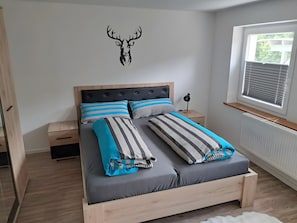 Een slaapkamer met een bed, gestreepte kussens, een houten hoofdbord, een nachtkastje, een aan de muur bevestigde decoratie van een hertenhoofd en een raam met jaloezieën.