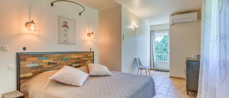 A bedroom with a wooden headboard, two pillows, a bed, a nightstand, a chair, a desk, an air conditioning unit, and a window with a view of greenery.