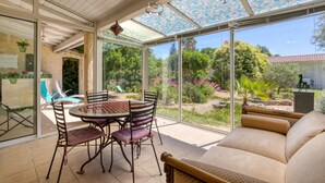 A sunlit patio with a table and chairs, a cushioned sofa, and a view of a garden through large glass doors. - Gîte du midi - gîtes domaine la noria (Cazouls-lès-Béziers)
