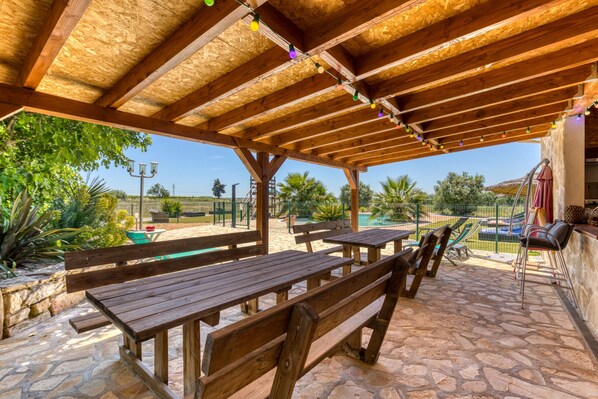 An outdoor patio with wooden benches and tables, a stone floor, and a wooden pergola with string lights.