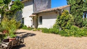 A rustic house with a gravel driveway, a wooden cart, and a variety of plants. - Gîte l'écluse - Domaine La Noria Holiday Homes (Cazouls-lès-Béziers)