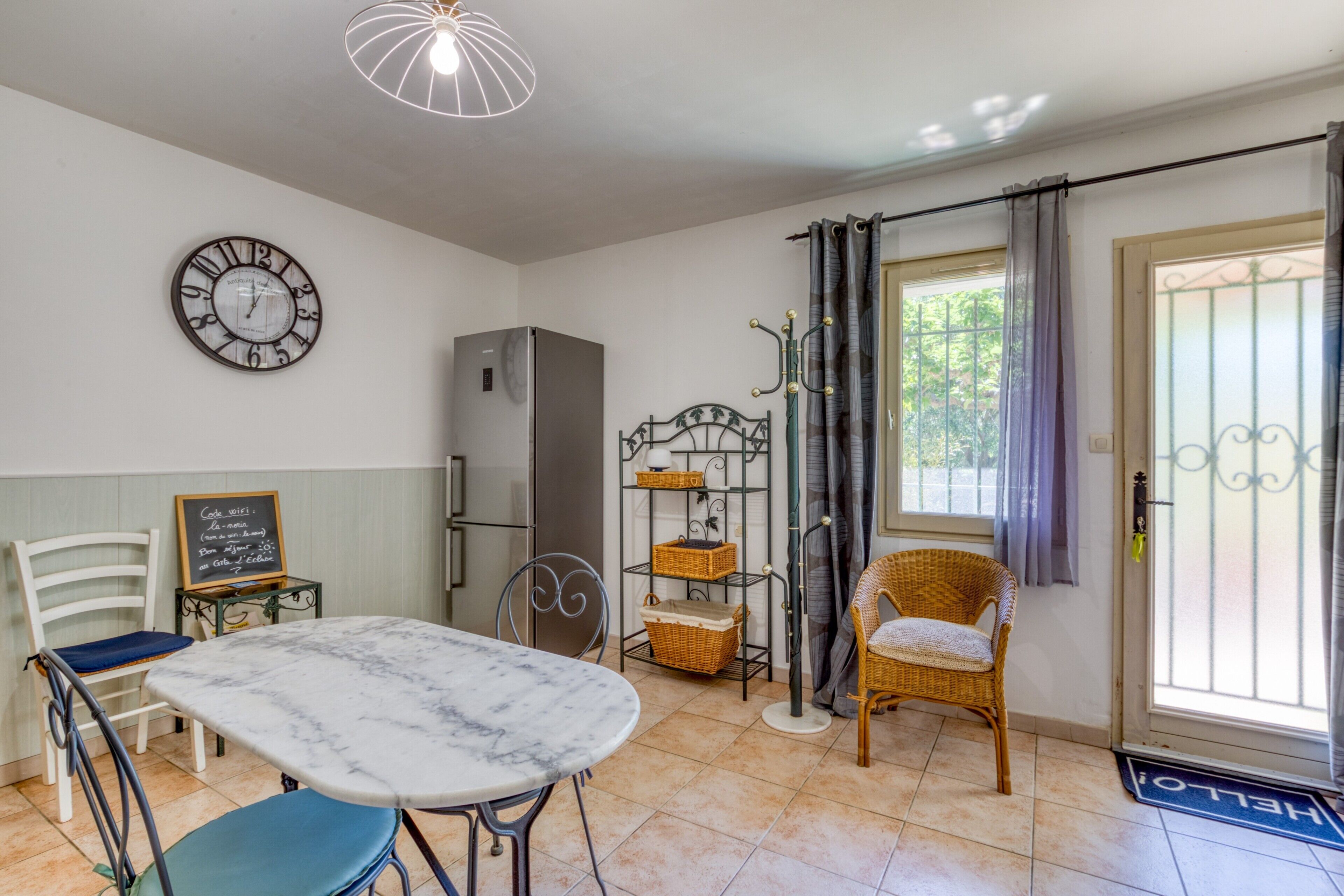 A kitchen with a marble table, chairs, a refrigerator, a clock, wicker baskets, and a door with a doormat.
