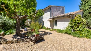 A rustic building with a gravel driveway, a wooden cart with a potted plant, and a large tree in the foreground. - Gîte l'écluse - Domaine La Noria Holiday Homes (Cazouls-lès-Béziers)