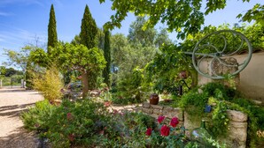 A garden with a stone wall, a large metal wheel, and various plants and flowers. - Gîte l'écluse - Domaine La Noria Holiday Homes (Cazouls-lès-Béziers)