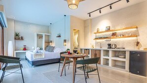 Una habitación de hotel moderna con cama, una mesa de comedor con sillas, una zona de cocina con refrigerador y un baño visible a través de una puerta abierta.
