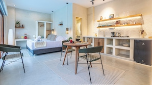 Una habitación de hotel moderna con cama, zona de comedor con mesa y sillas, cocina con refrigerador y estantes, y baño con ducha e inodoro.