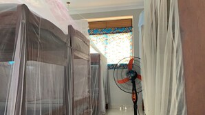 Une chambre avec un lit recouvert d’une moustiquaire, un ventilateur sur pied et une fenêtre ornée de rideaux à motifs.