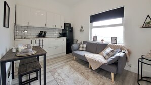 A compact living space with a kitchenette, a grey sofa, a small table with a candle, and a window with blinds. - Modern City Apartment in London (London)