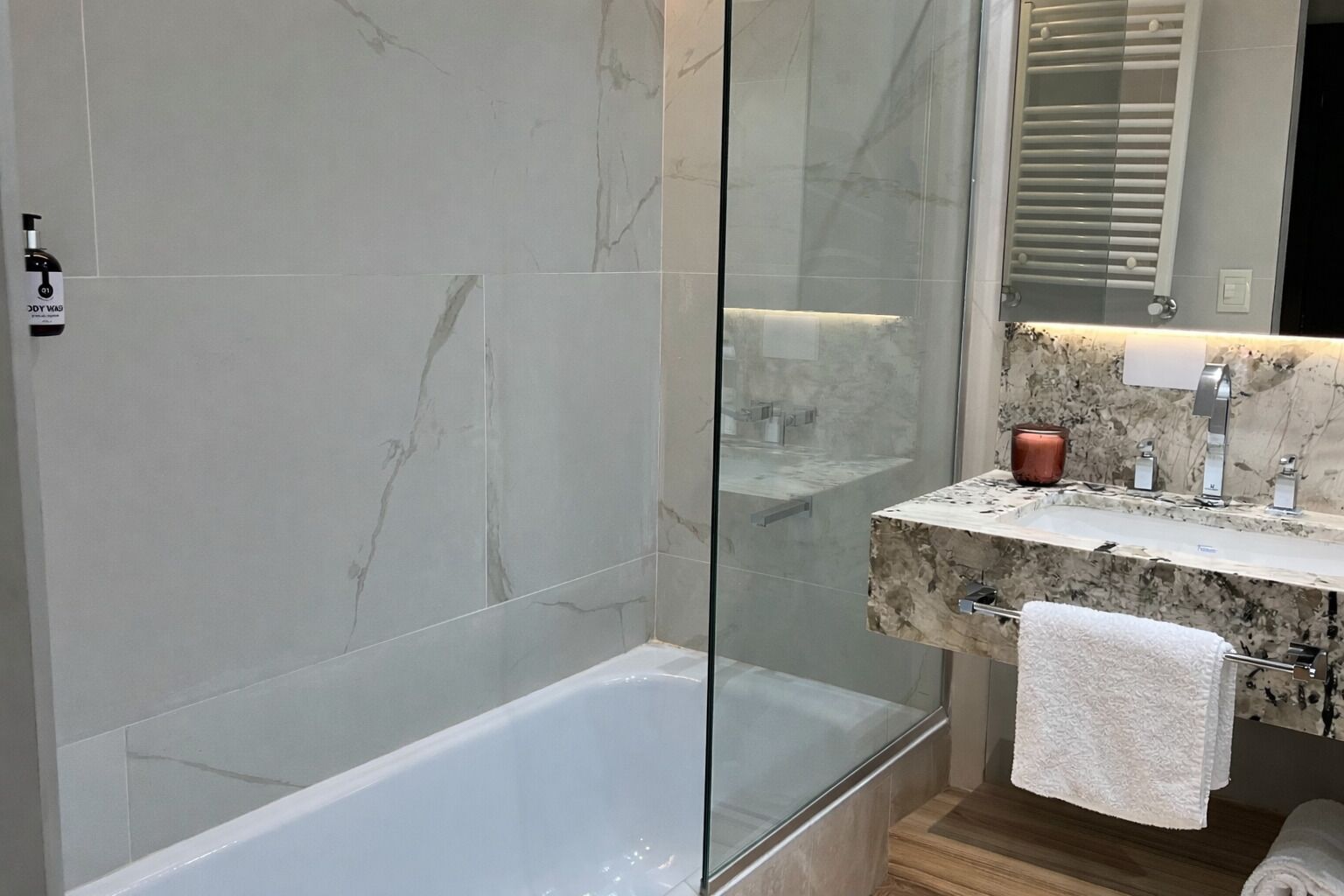 Un baño moderno con ducha envuelta en vidrio, pared con azulejos de mármol, piso de madera y encimera de mármol con un candelabro de cobre.