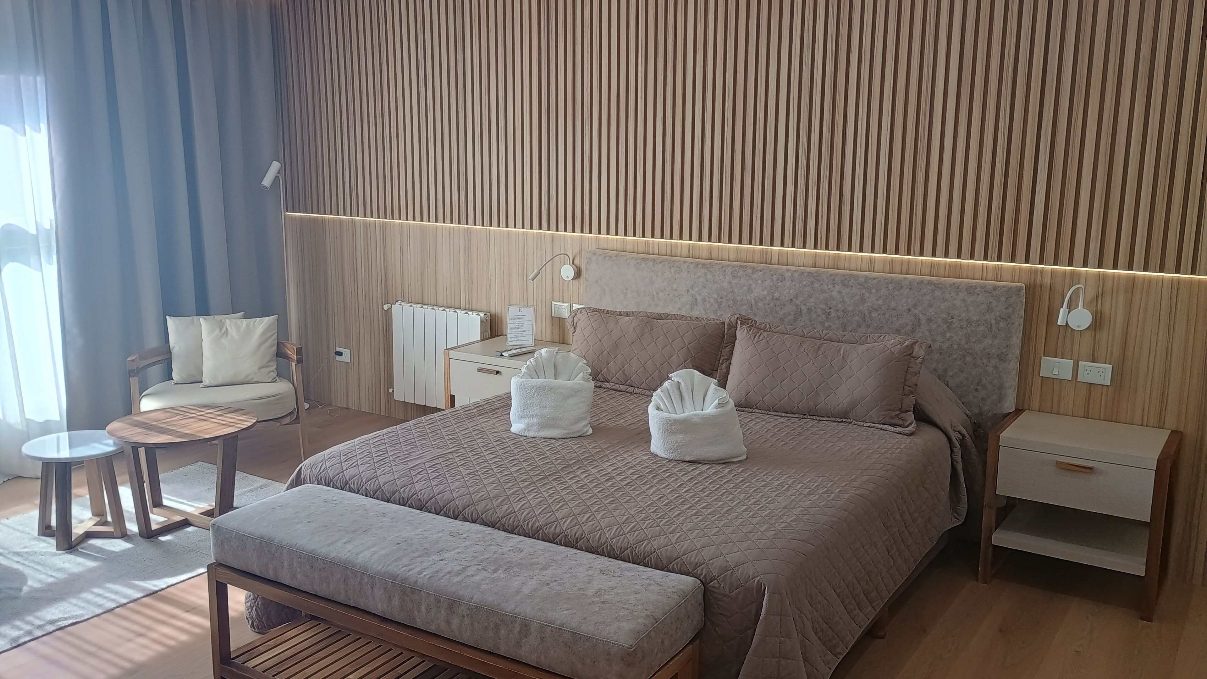 Habitación de hotel moderna con una cama grande, mesitas de noche, una zona de estar con una silla y una pequeña mesa, y suelo de madera.