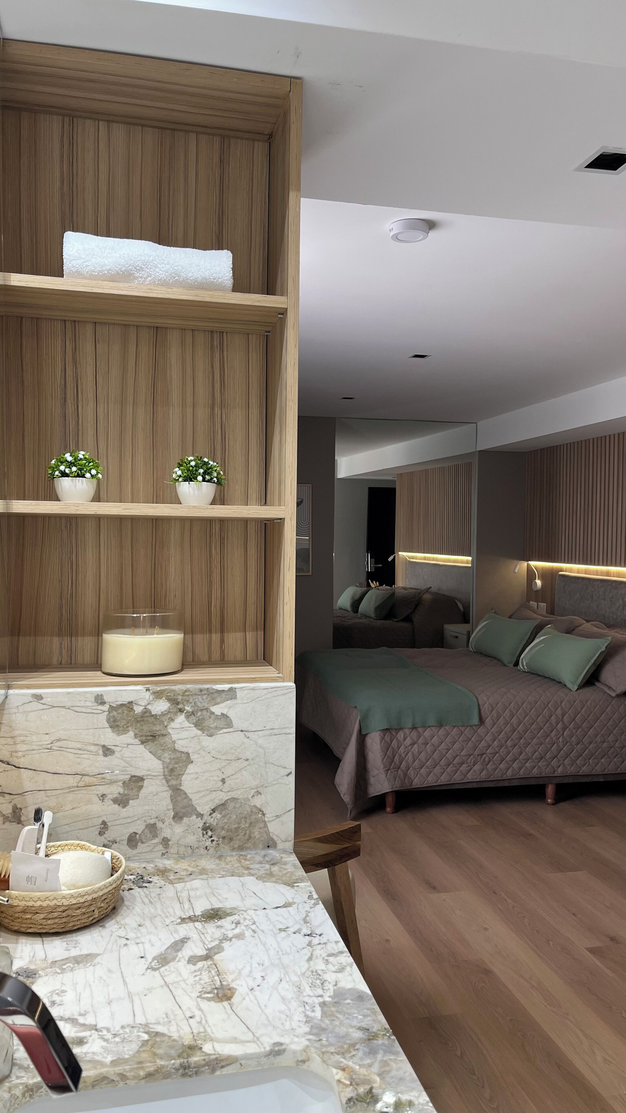 A modern bedroom with a wooden headboard, a bed with a quilted cover, bedside lamps, and a small potted plant on a shelf.