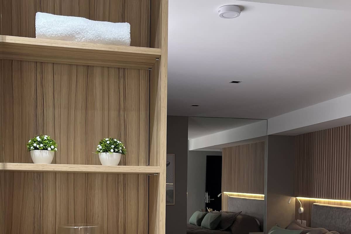 A modern bedroom with a wooden headboard, a bed with a quilted cover, bedside lamps, and a small potted plant on a shelf.