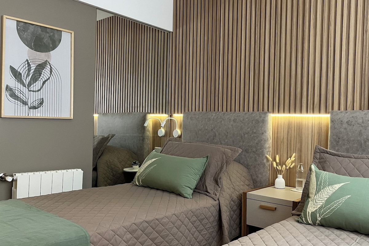 A modern bedroom with two beds, a nightstand, a bedside table, and a wall-mounted lamp.