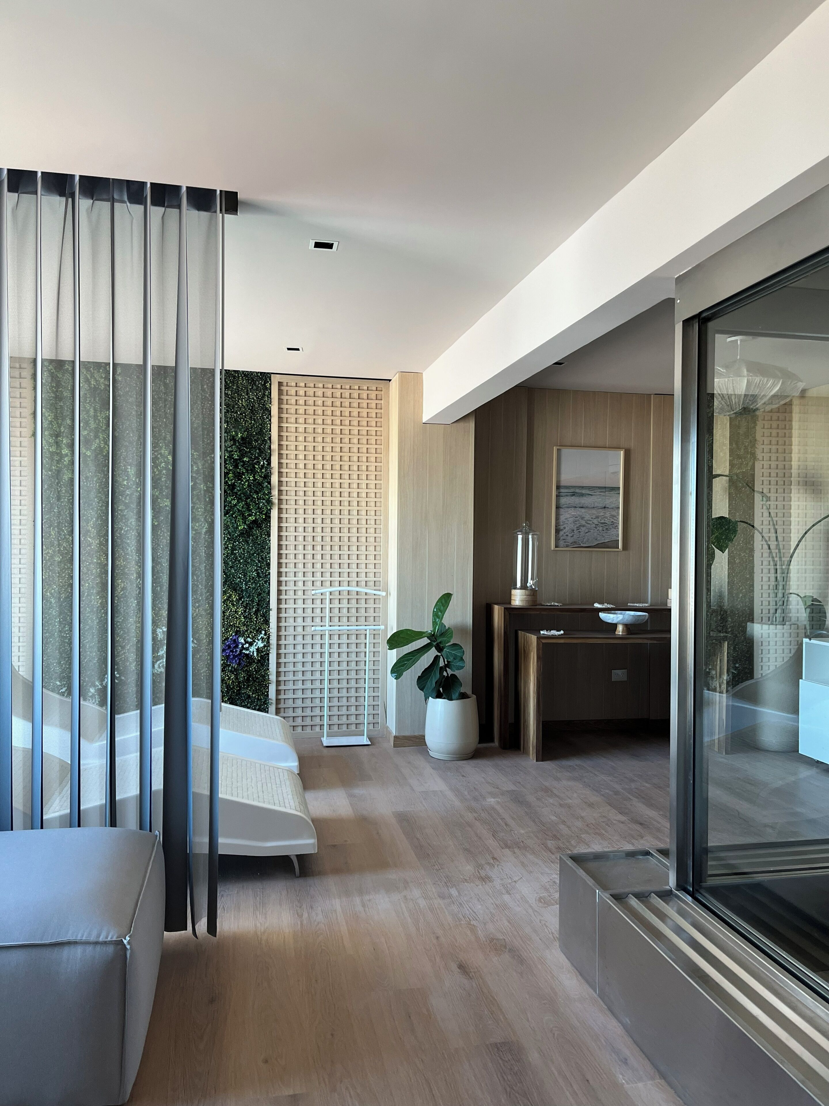 Habitación de hotel moderna con balcón, un amplio ventanal con cortinas, suelo de madera, una pequeña planta y vistas al mar.