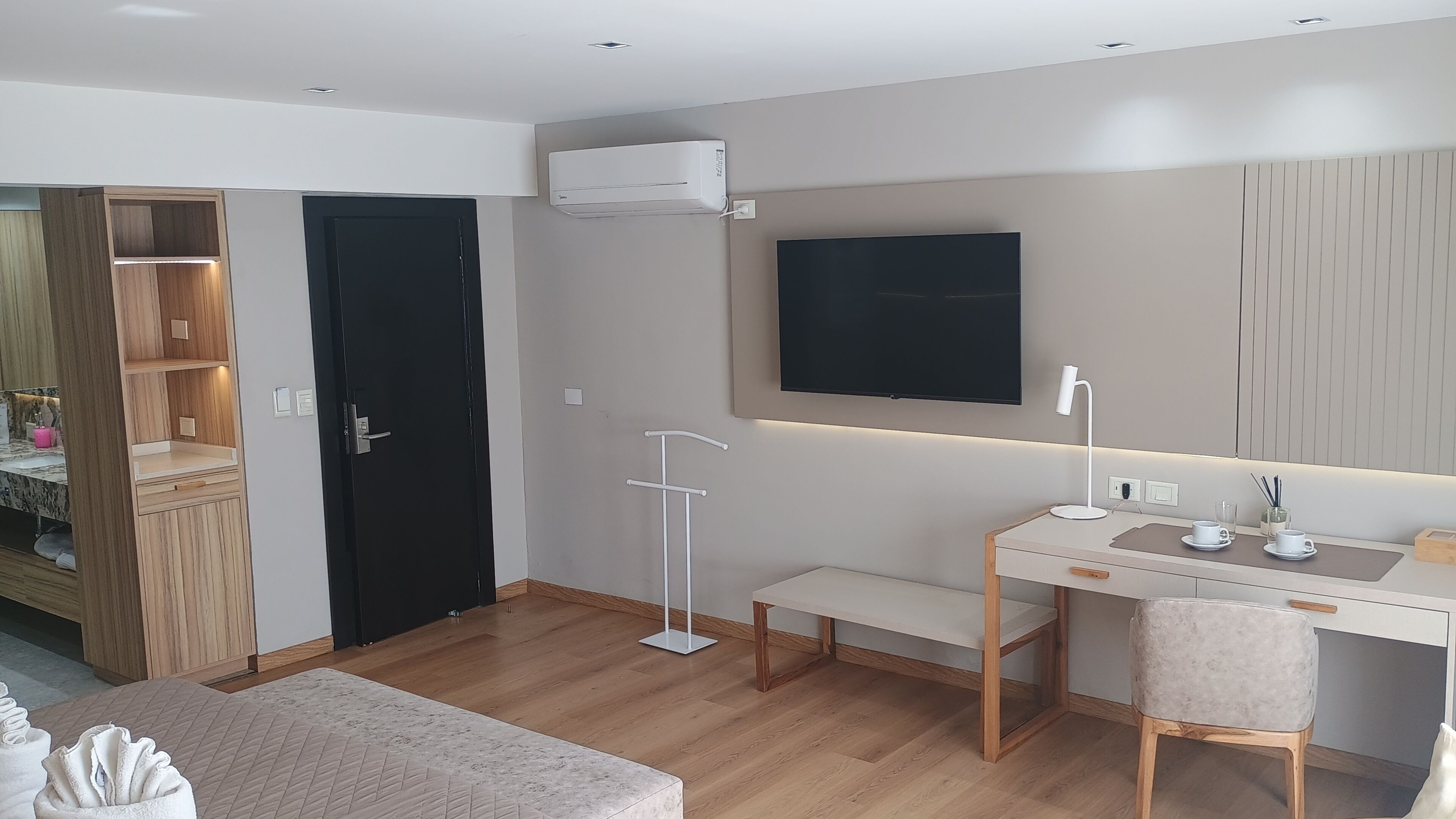 Una habitación de hotel moderna con un armario de madera, una puerta negra, un televisor de pantalla plana montado en la pared, un escritorio con una silla y una mesita de noche con una lámpara.