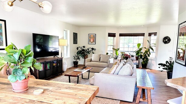 A spacious living room with a wooden dining table, a sofa, and a TV. - Stay between Glacier and Whitefish-cozy stylish (Columbia Falls)