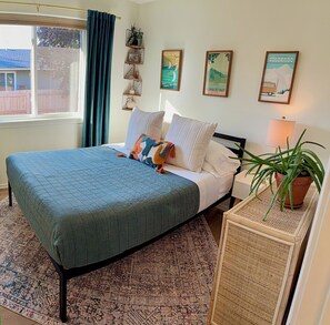 Un dormitorio con cama, almohadas, una mesita de noche con lámpara, una planta en maceta y decoración en la pared.