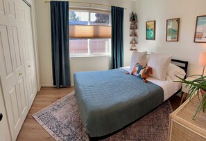 Un dormitorio con cama, ventana con cortinas, mesita de noche con lámpara y adornos en la pared.