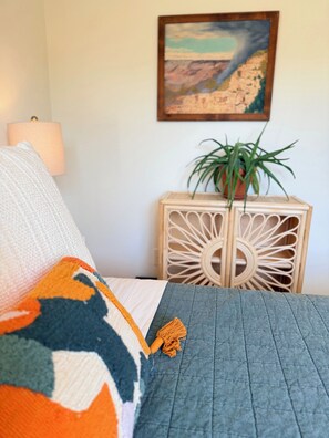 Een bed met een gebloemd dekbed, een kussen, een wandlamp, een ingelijste schilderij en een houten nachtkastje met een plantje.