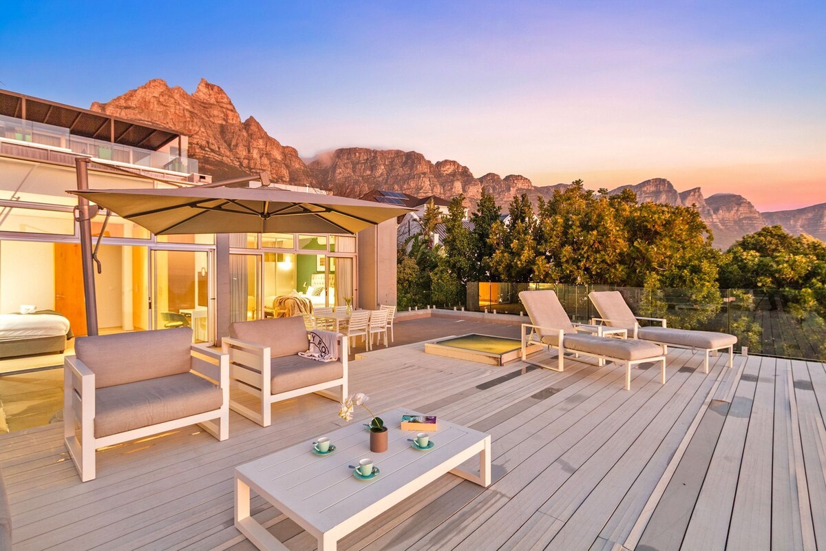 A modern outdoor deck with lounge chairs, a table, and a glass-enclosed room with a view of mountains.