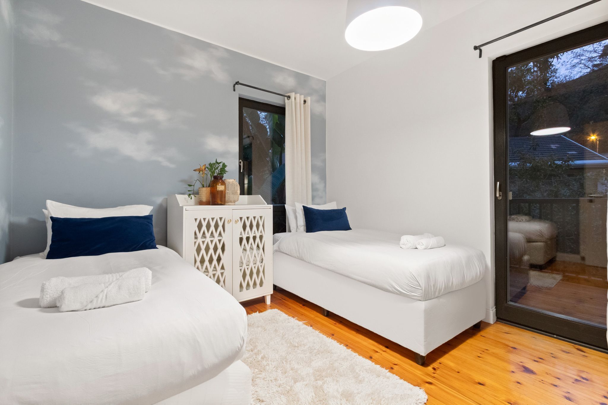 A modern bedroom with two single beds, a white wardrobe, and a large sliding glass door leading to an outdoor area.