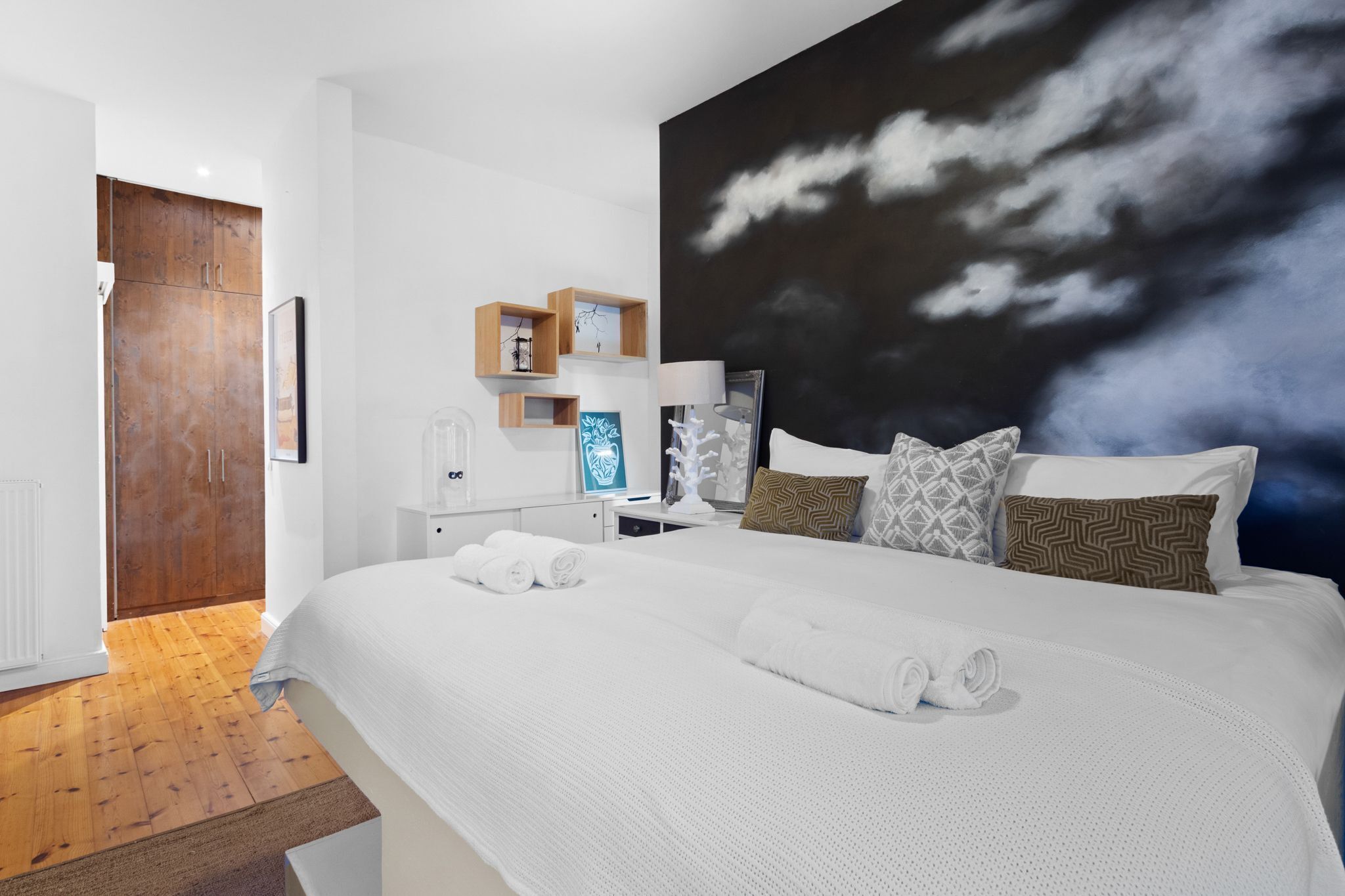A modern hotel room with a large bed, a dark cloud-themed wall mural, wooden cabinets, and a wooden floor.