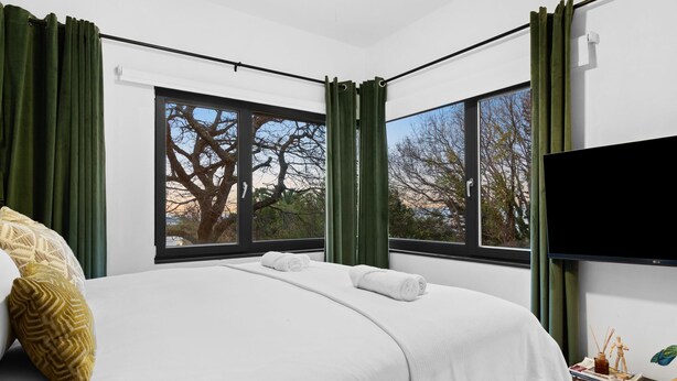 A modern hotel room with a large bed, green curtains, a flat-screen TV, and a view of trees through the windows. - Higgo Crescent Villa (Cape Town)