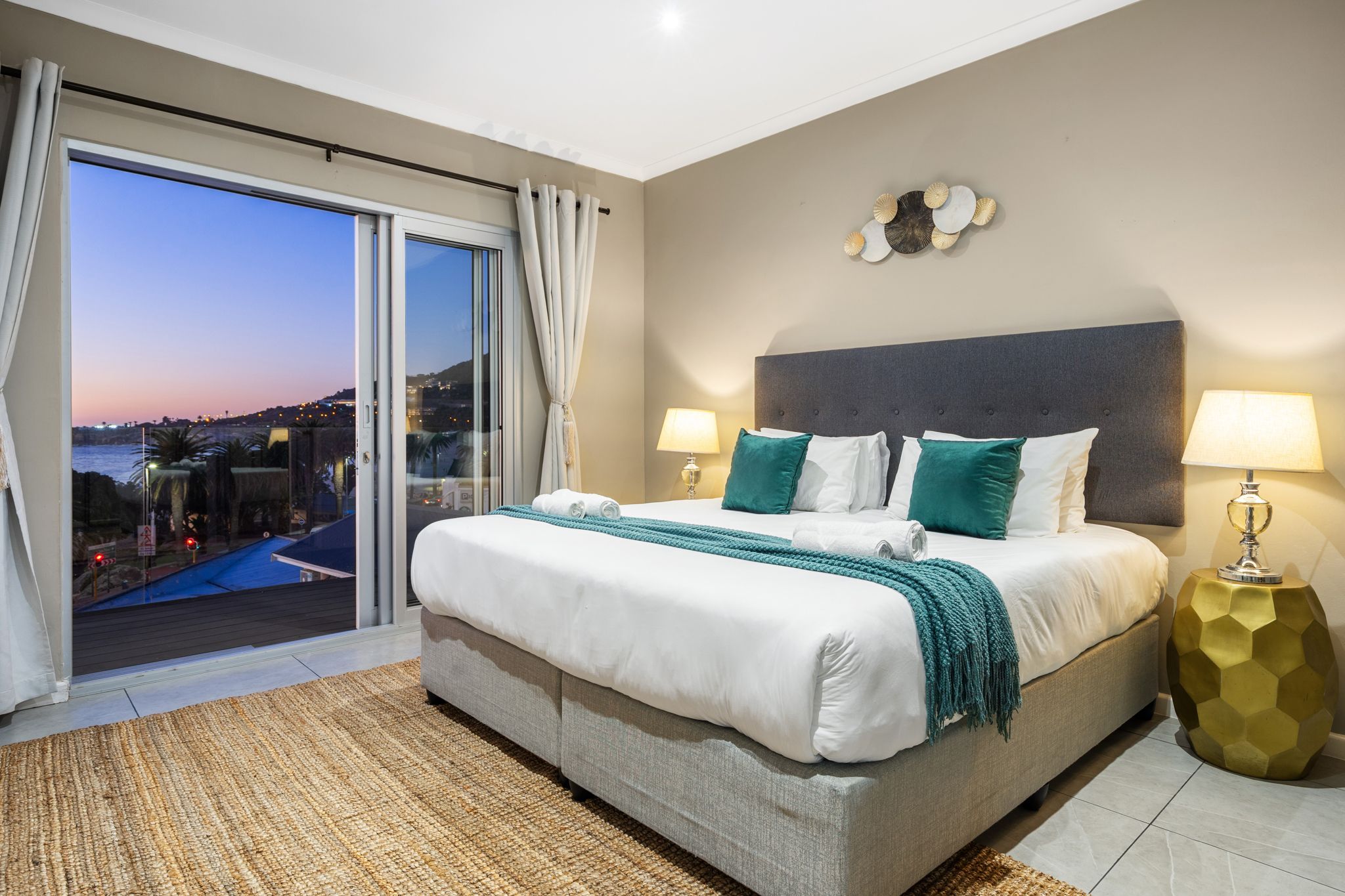 A bedroom with a large bed, two bedside lamps, a view of a pool, and a cityscape at dusk.