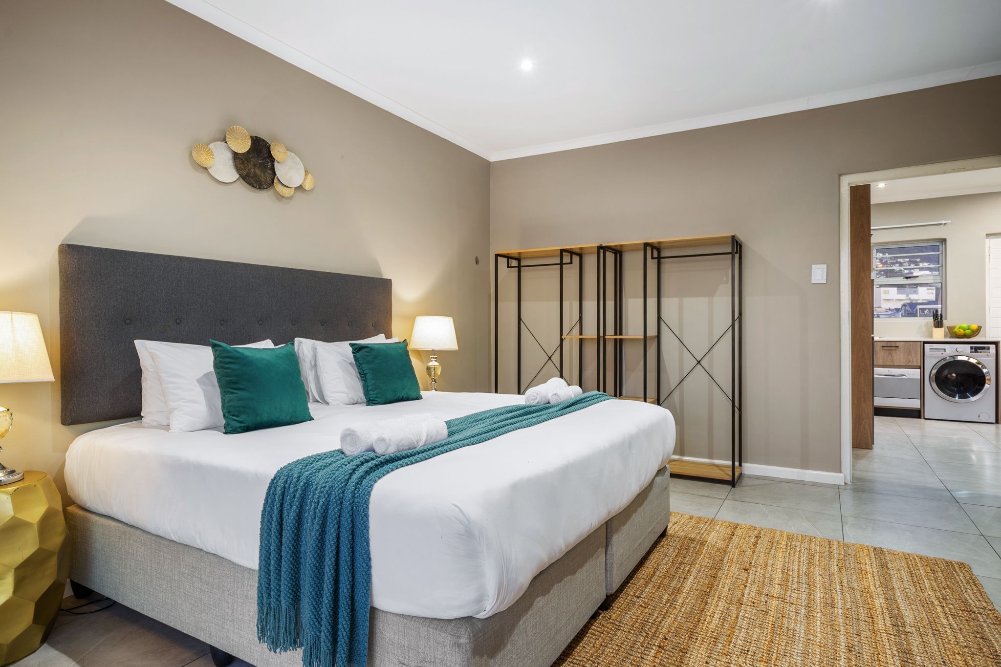 A neatly made bed with white and teal bedding, a grey headboard, and a woven rug. A small table with a lamp is beside the bed.