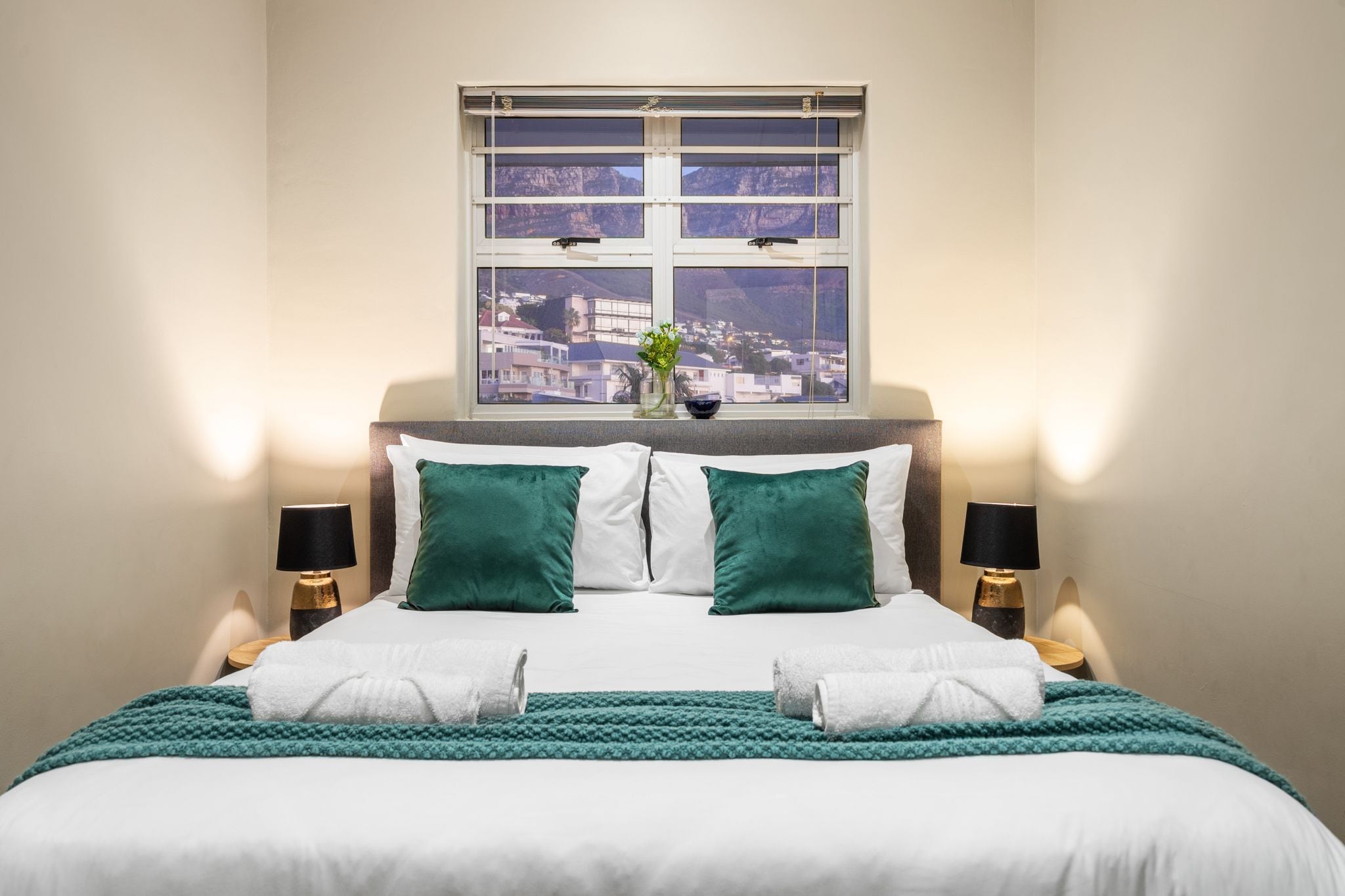 A neatly made bed with white and green bedding, two lamps on either side, and a window with a view of buildings and mountains.