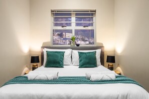 A neatly made bed with white and green bedding, two lamps on either side, and a window with a view of buildings and mountains.