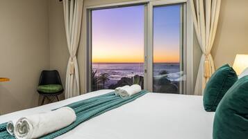 A hotel room with a bed, a view of the ocean at sunset, a green chair, and a teal throw blanket.