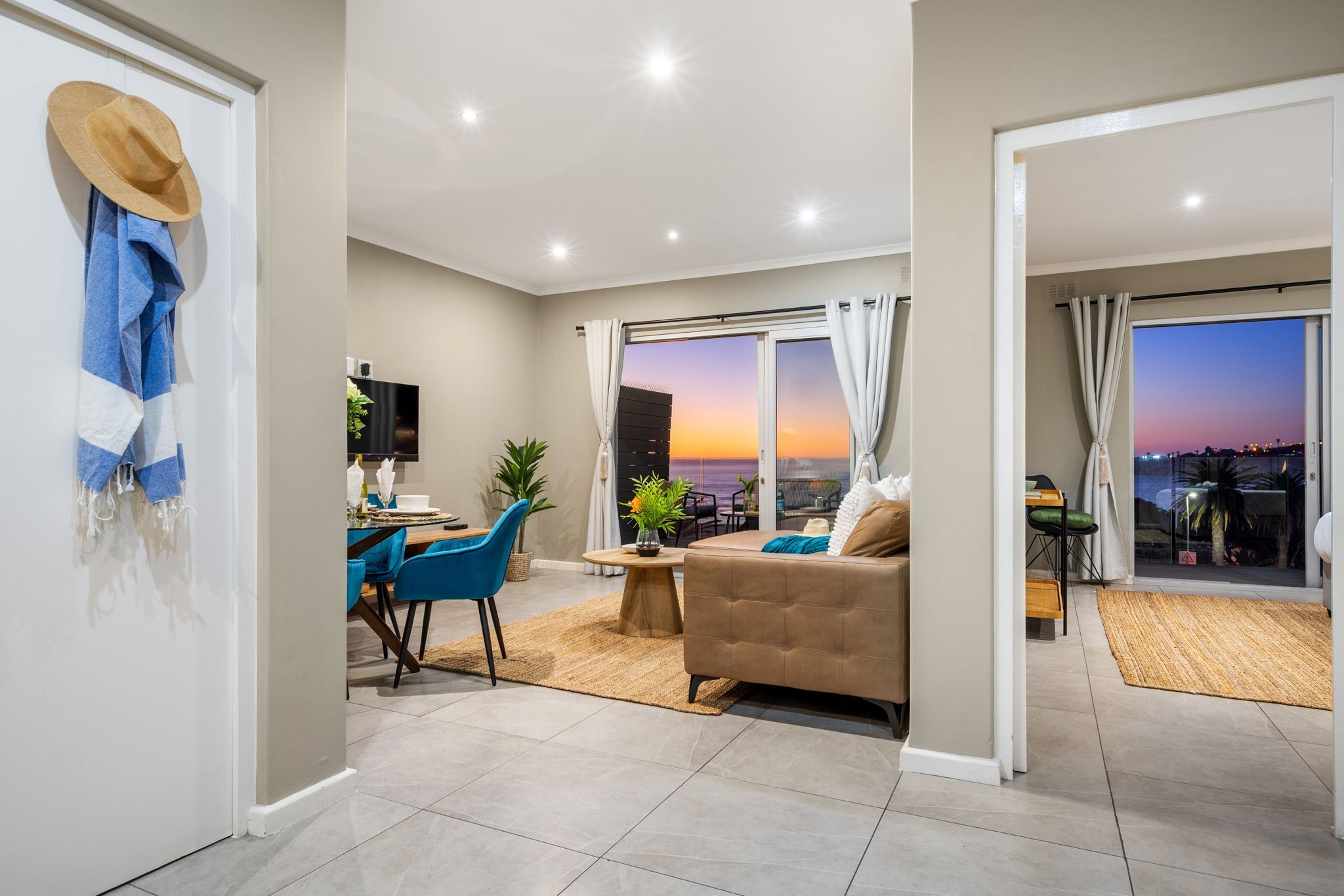 A modern living room with a sofa, dining area, and a view of the sunset through large windows.