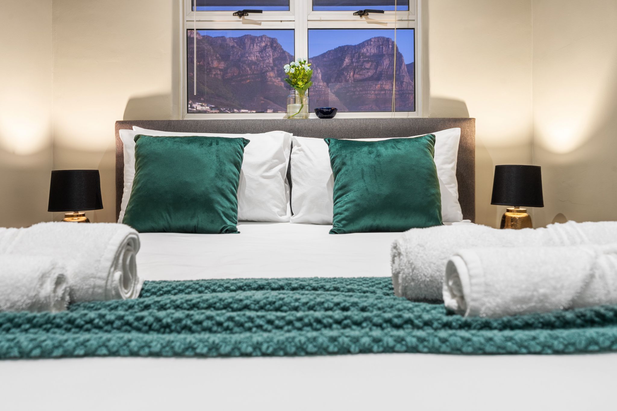 Een net opgemaakt bed met groene en witte kussens, uitzicht op bergen door een raam, en een smaragdgroen vloerkleed.