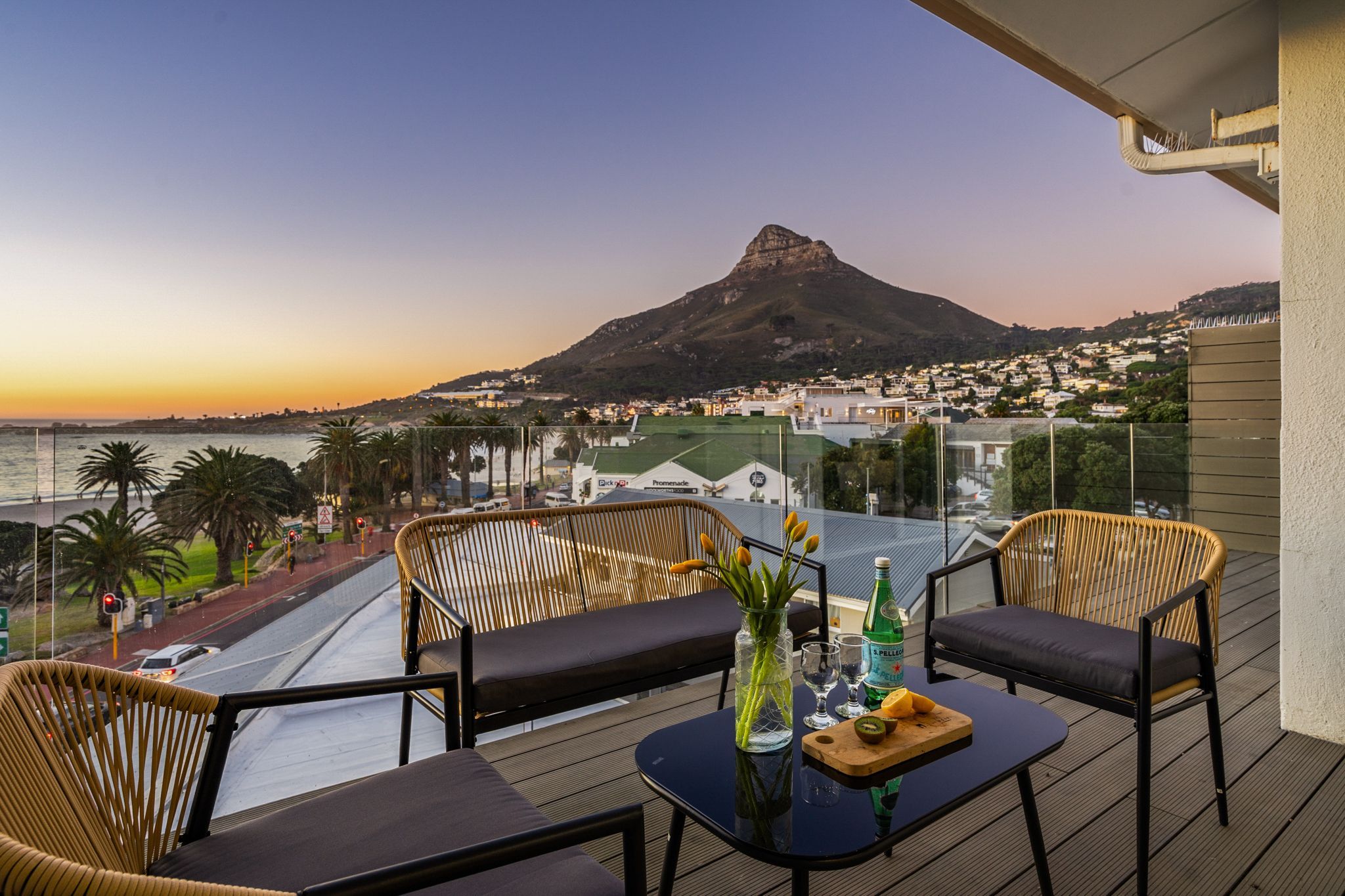 Un balcon avec des fauteuils en osier, une table avec une bouteille et des verres, et une vue sur une montagne et une ville.