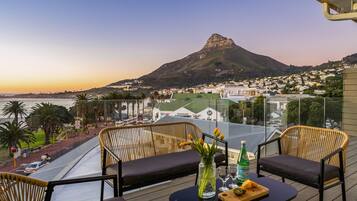 A balcony with wicker chairs, a table with a bottle and glasses, and a view of a mountain and town.