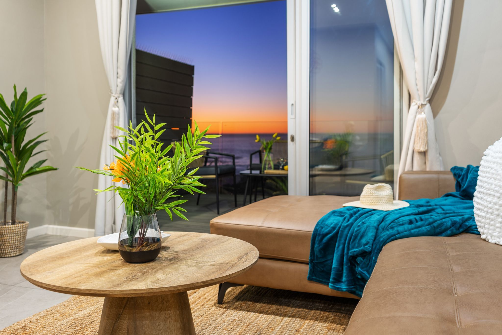 A modern living room with a round wooden table, a sofa, and a view of the sunset through a glass door.