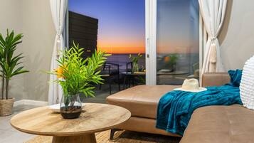 A modern living room with a round wooden table, a sofa, and a view of the sunset through a glass door.