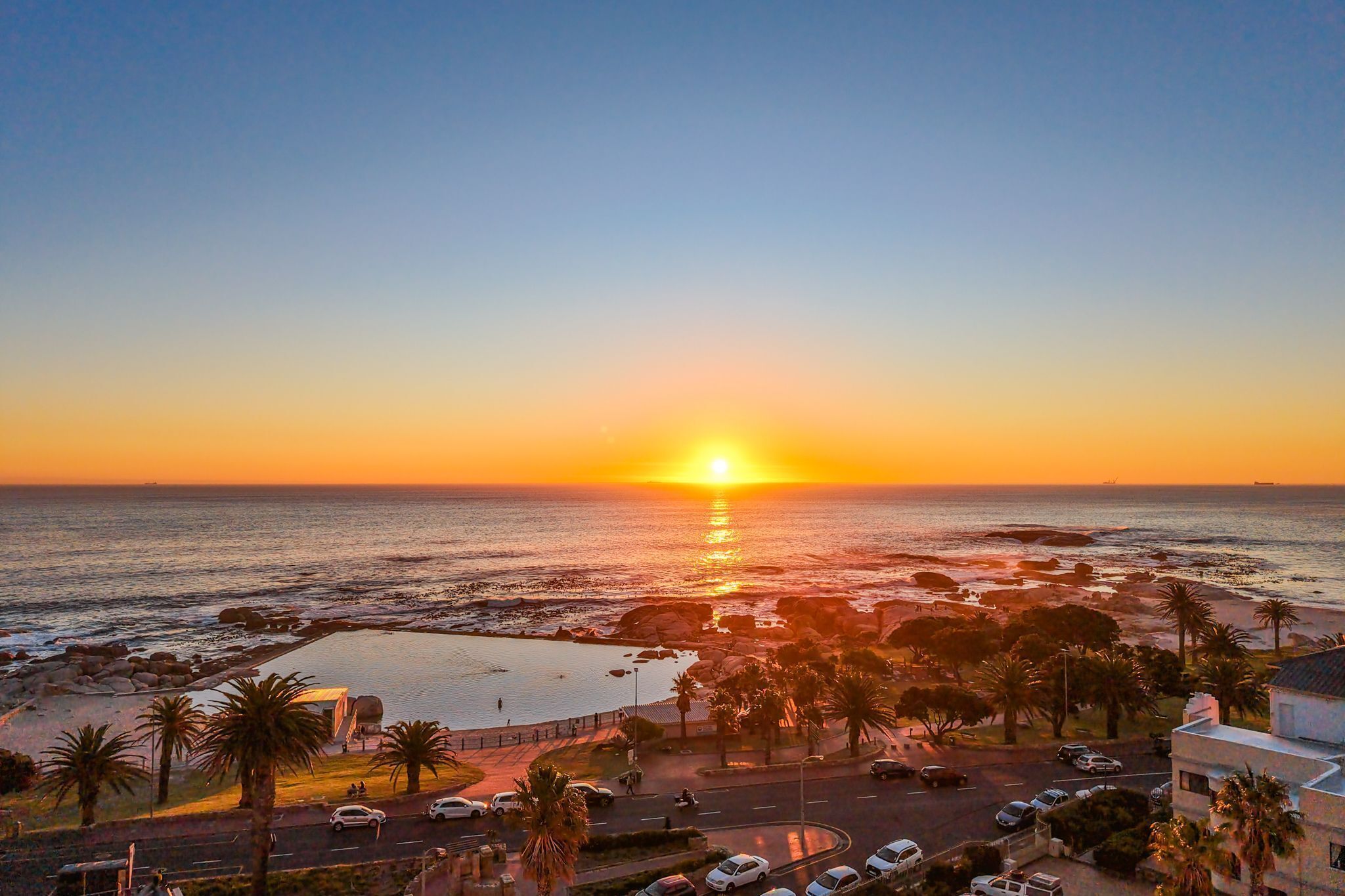 A coastal sunset view with a parking area, palm trees, and a road leading to the beach.