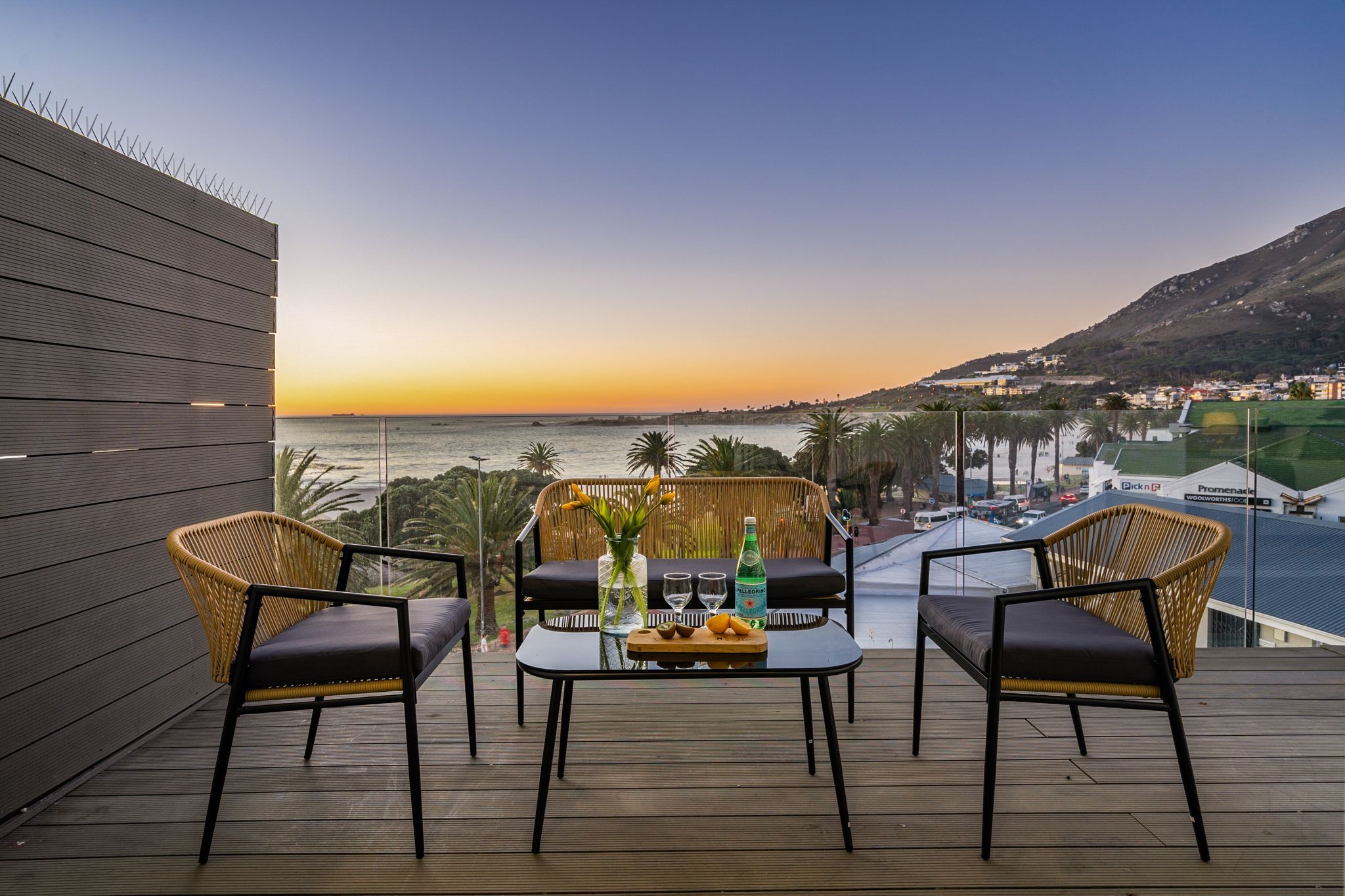 A balcony with wicker chairs, a table with drinks, and a view of the sea and hills.