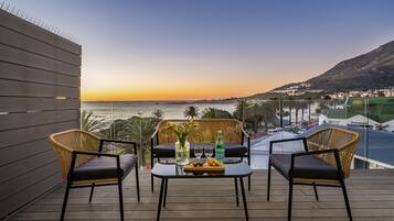 A balcony with wicker chairs, a table with drinks, and a view of the sea and hills.