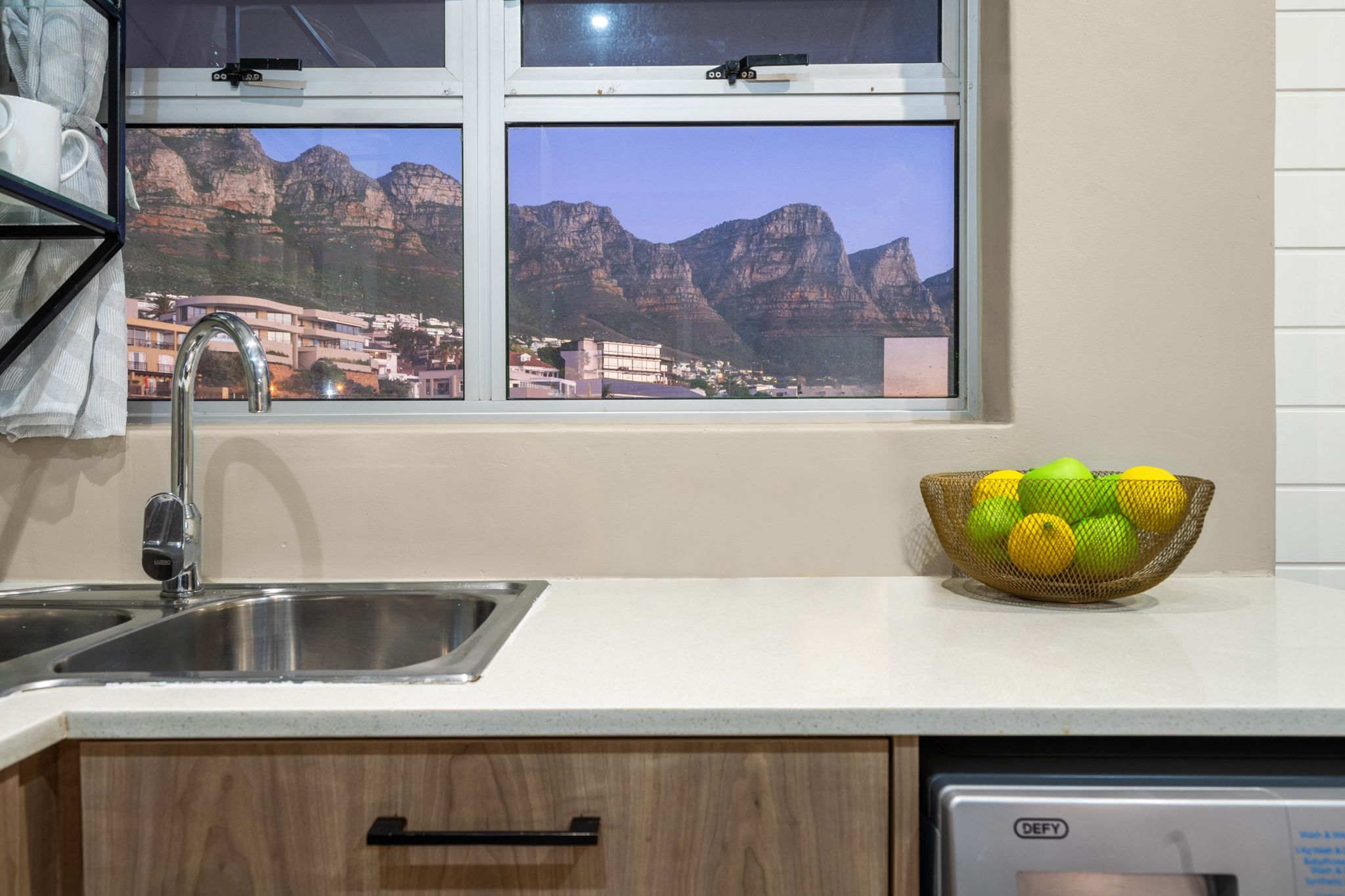 A kitchen with a stainless steel sink, a bowl of fruit on the counter, and a window with a view of mountains and buildings.