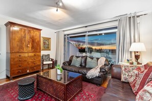 A living room with a brown leather sofa, a wooden cabinet, a glass coffee table, and a view of a sunset through large windows. - Firdale Mews 5 (Cape Town)