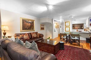 A living room with a brown leather sofa, a glass coffee table, and a red patterned rug. There is a dining area with a table and chairs, and a kitchen area with a window and a counter. - Firdale Mews 5 (Cape Town)