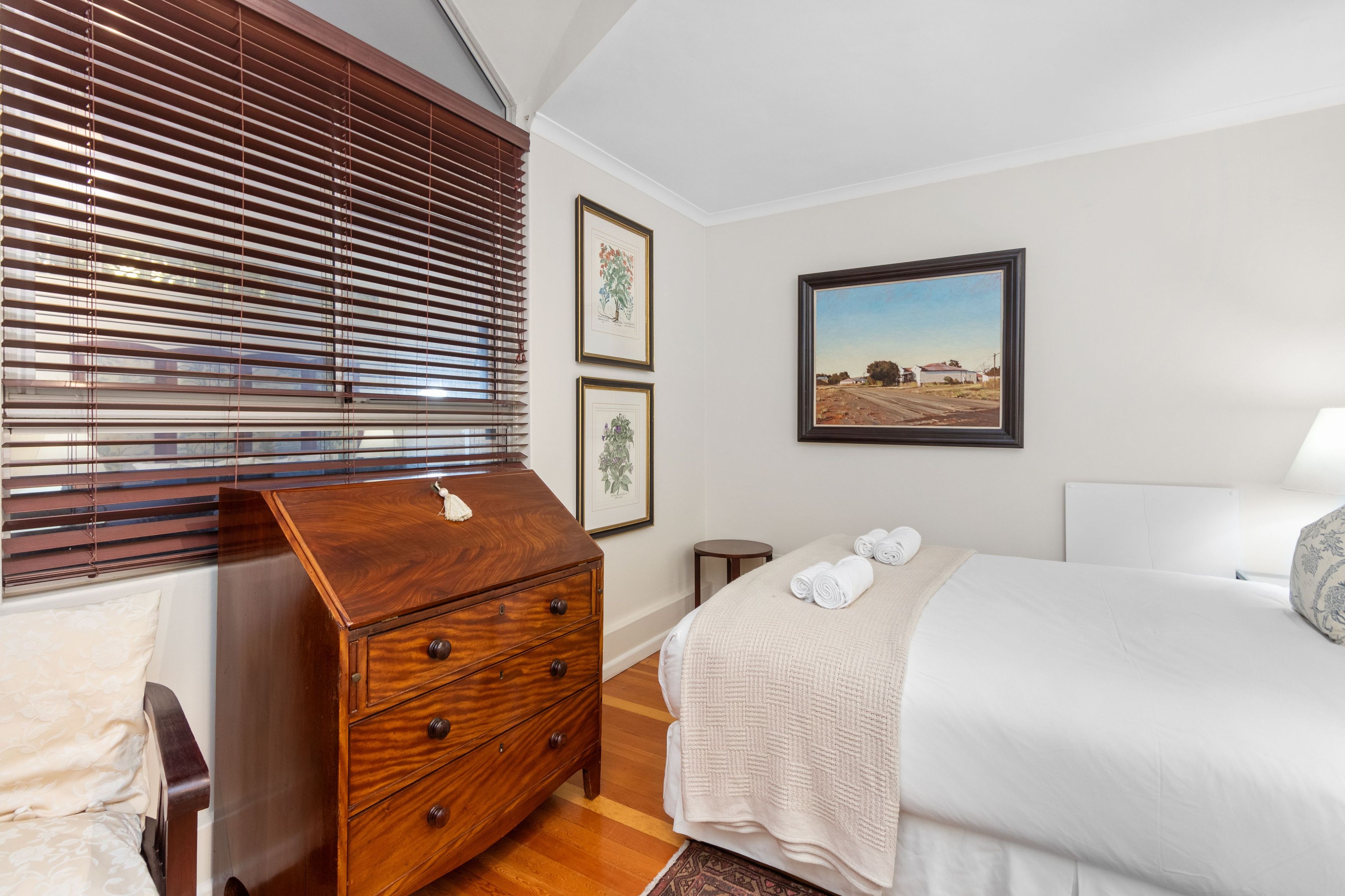 Een slaapkamer met een houten dressoir, een bed met wit beddengoed, een lijst met een landschap en een raam met jaloezieën.