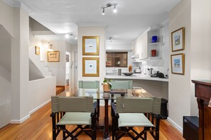 A dining area with a glass table and chairs, a kitchen in the background, and framed pictures on the walls. - Firdale Mews 5 (Cape Town)