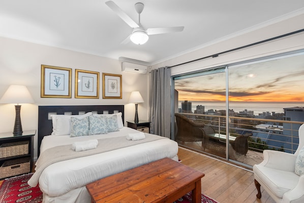 A modern bedroom with a large bed, a ceiling fan, and a city view through floor-to-ceiling windows. - Firdale Mews 5 (Cape Town)