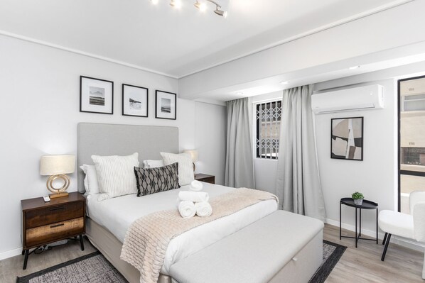 A modern hotel room with a large bed, a bedside table with a lamp, a small side table with a plant, and a window with curtains.